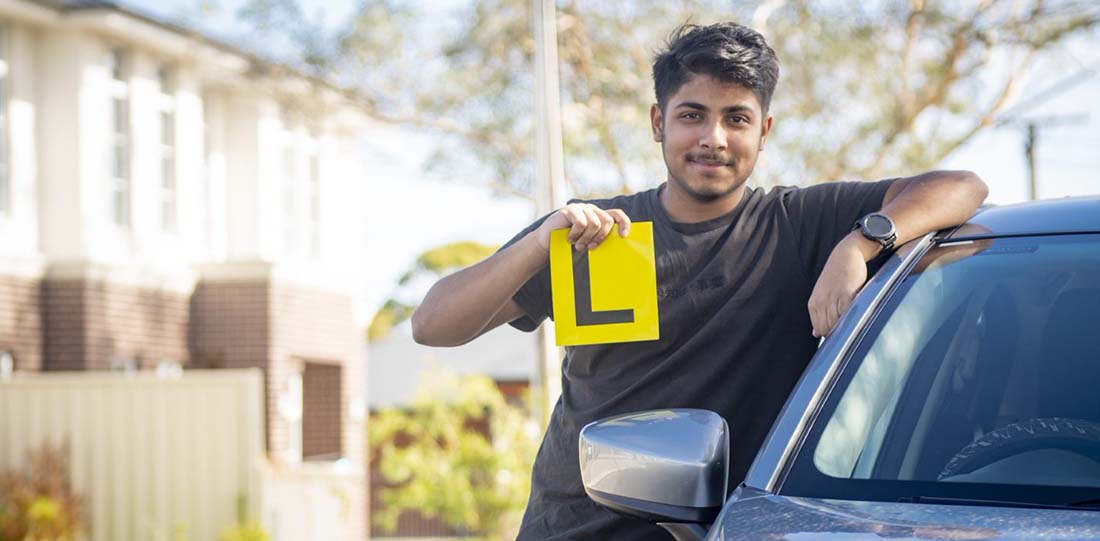 A young man leaning on his car holding a learners plate in his right hand